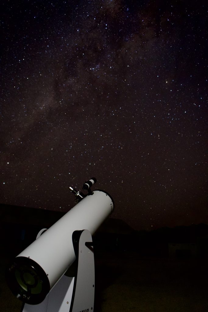 A white Dobsonian telescope points towards a dark night sky filled with countless stars and the faint glow of the Milky Way galaxy.